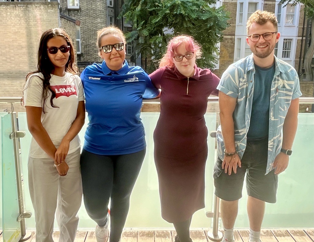 A picture of the Young Voices Sports team smiling at the camera, standing side by side in front of a glass railing with a building and trees in the background.