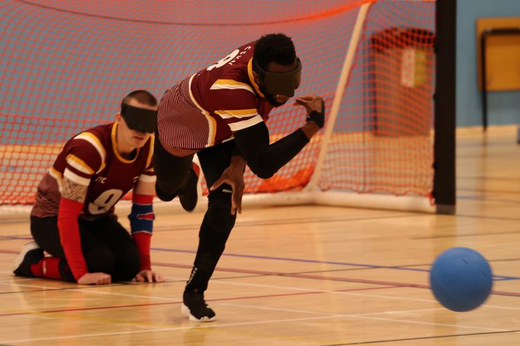Two male students are playing goalball in a sports hall, wearing maroon jerseys with white stripes. One student is in a kneeling position and the other is preparing to throw the ball.