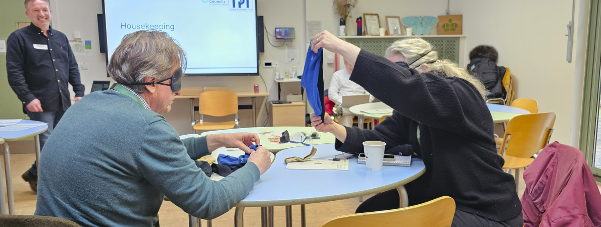 In a classroom setting, two individuals sit at a table with a projector screen in the background displaying the TPT Logo. The man on the left wears a blue shirt and a eye cover. The woman next to him is also wearing an eye cover but her face is obscured as she holds up another eye cover in front of her.