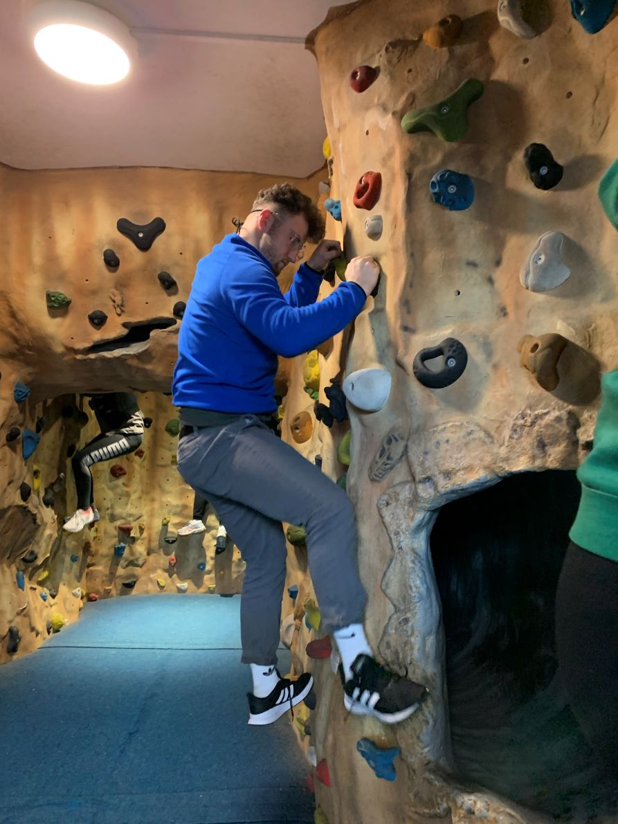 Tom is climbing up an indoor rock wall, wearing a blue jacket and grey trousers.