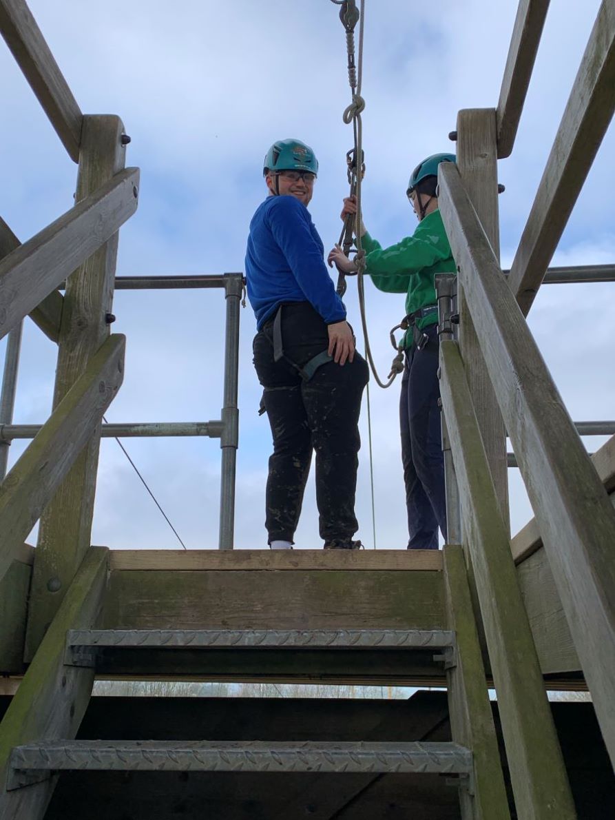 Tom is standing on a wooden platform, about to go down a zipline, being helped into a harness.