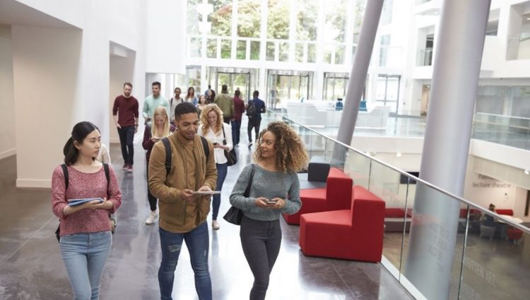 A group of students walking through a modern bright hallway, holding notebooks and chatting as they navigate their way through the space.