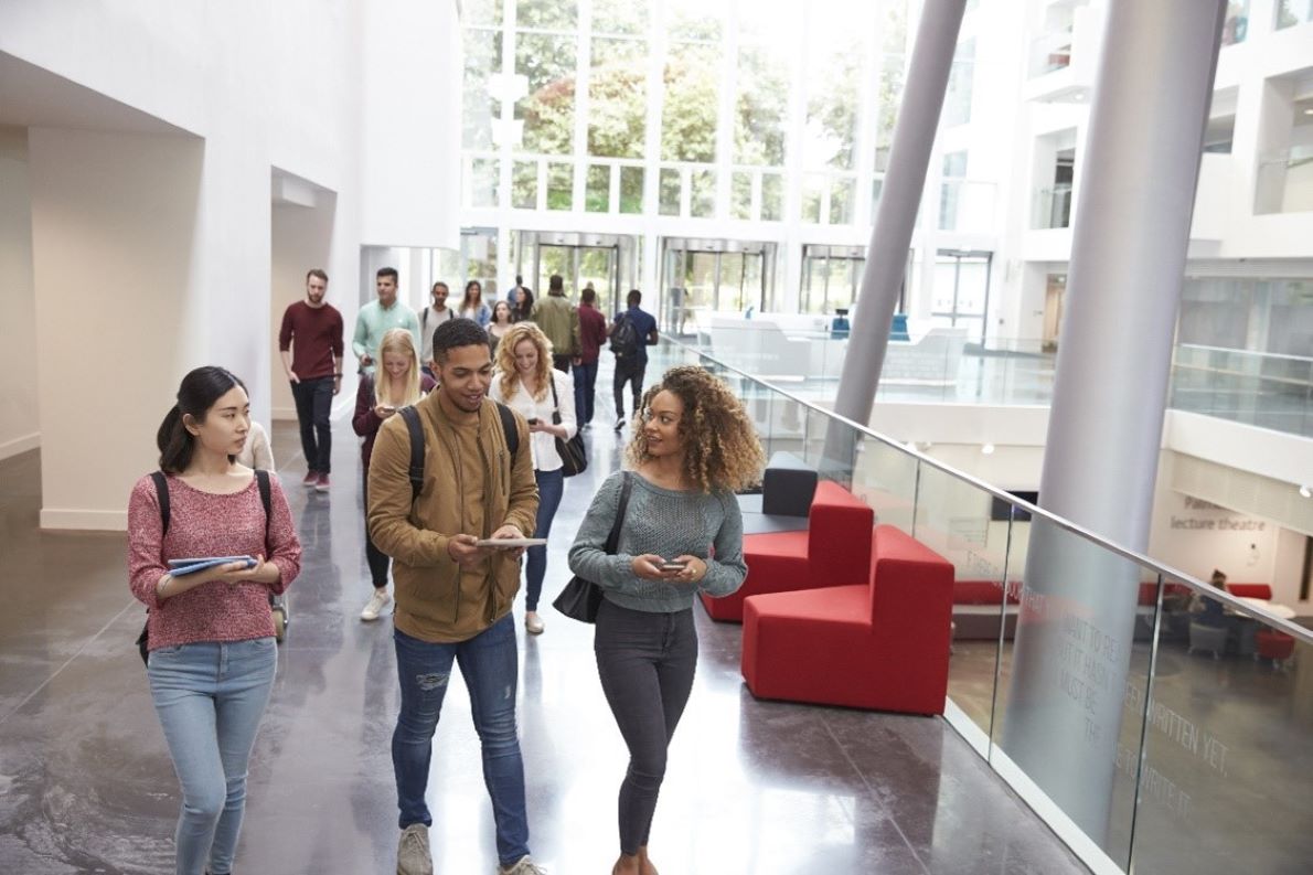 A group of students walking through a modern bright hallway, holding notebooks and chatting as they navigate their way through the space.
