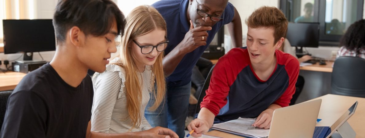 A teacher is supporting three young students with their work, deep in discussion and engaged in learning in a bright inclusive setting.