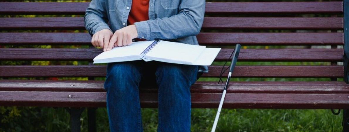 A student with a long cane is sitting on a bench reading a large open book in braille/