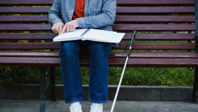 A student with a long cane is sitting on a bench reading a large open book in braille/