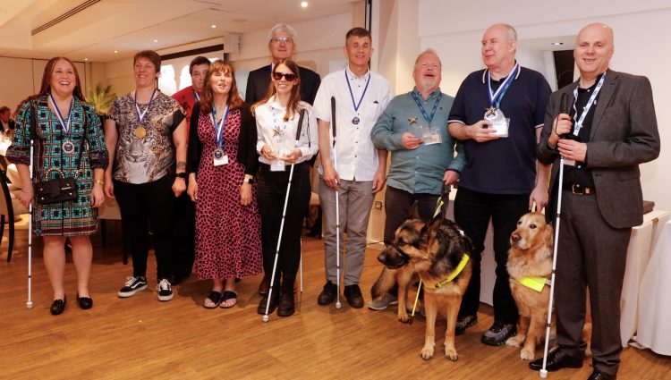 Sight Loss Council volunteers and Thomas Pocklington Trust staff stood in a line smiling at the camera at the Rodney Powell Awards for Volunteering where they won an award for their involvement in the development of the bus and coach driver training materials. Two guide dogs (an Alsatian and Labrador) are pictured in the photo.