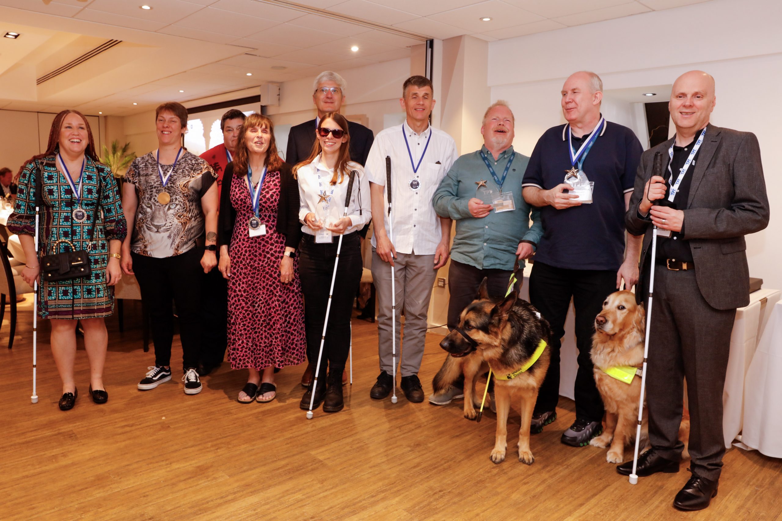 Sight Loss Council volunteers and Thomas Pocklington Trust staff stood in a line smiling at the camera at the Rodney Powell Awards for Volunteering where they won an award for their involvement in the development of the bus and coach driver training materials. Two guide dogs (an Alsatian and Labrador) are pictured in the photo. Caption: Sight Loss Council volunteers recently won a Rodney Powell Award for Volunteering for their involvement in the development of the training materials.