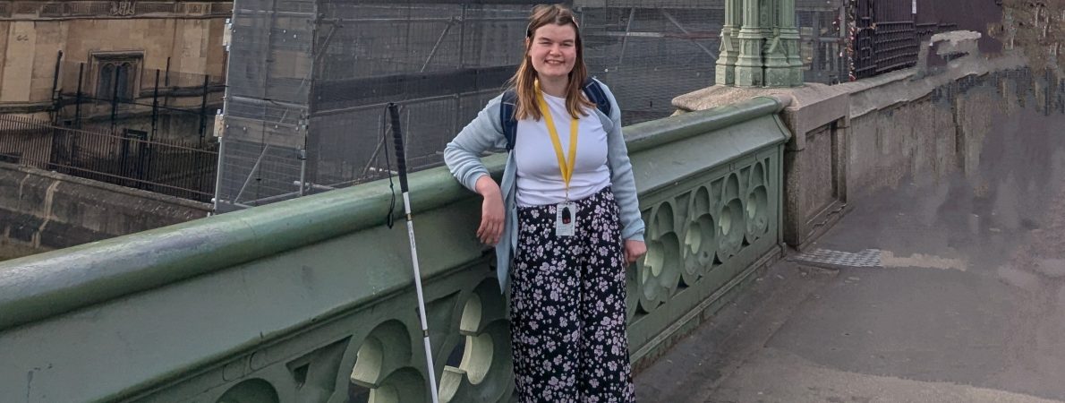 Sarah is standing on Westminster Bridge, holding her white cane and wearing patterned trousers, with Big Ben in the background.