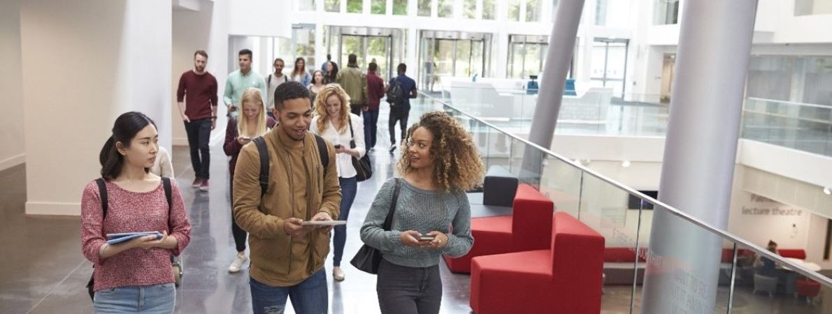 A group of students walking through a modern bright hallway, holding notebooks and chatting as they navigate their way through the space.