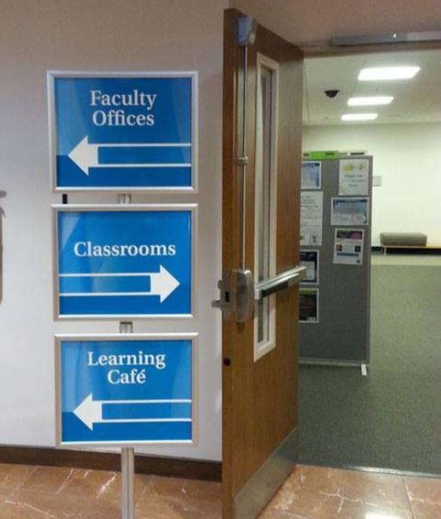 A wall with blue signs, pointing in different directions. Facility Offices points left, Classrooms points right, and Learning cafe points ;eft at the bottom.