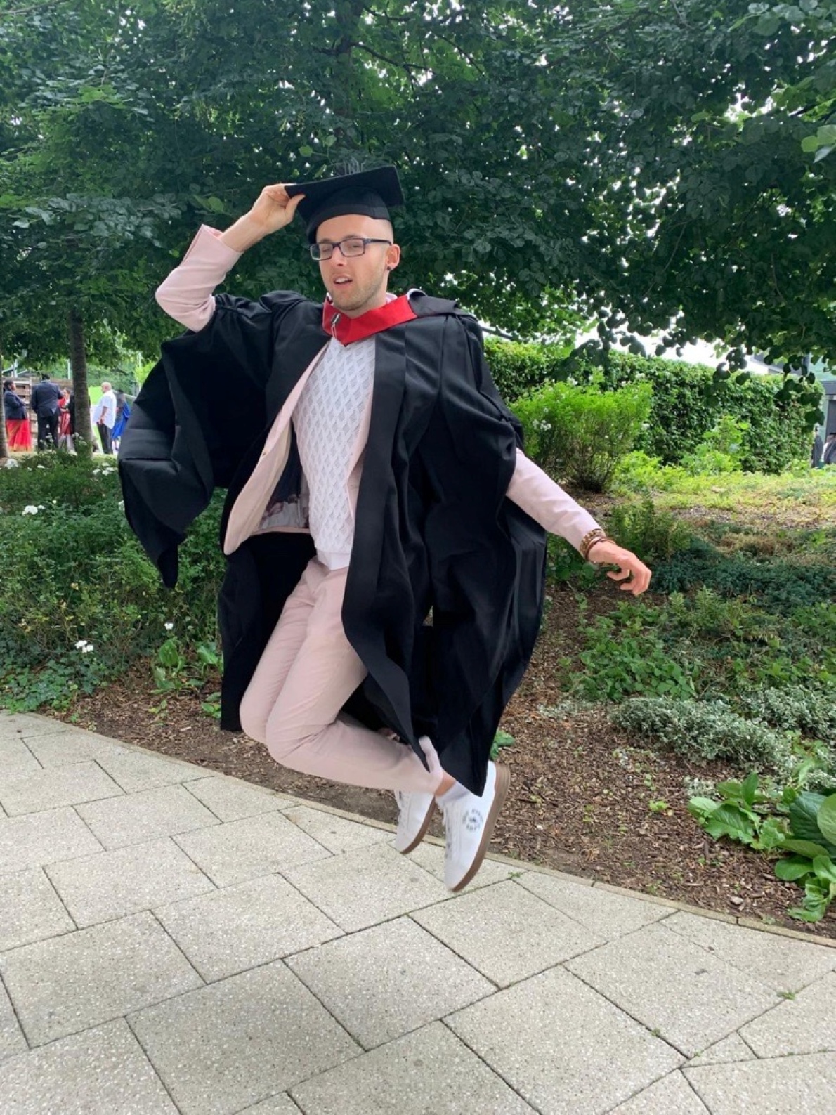Aleks is wearing his graduation gown and jumping in the air to celebrate, in an outdoor green space.