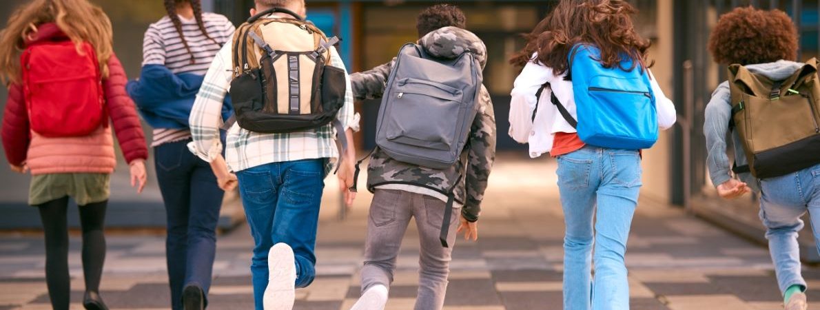 Five students wearing backpacks are walking towards a campus building.