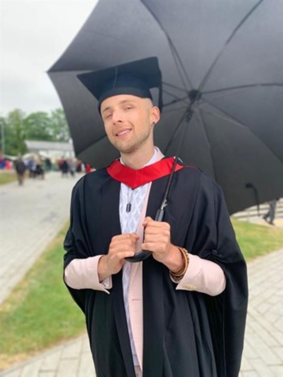Aleks is standing in a green space wearing his graduation gown and holding an black umbrella, smiling to the camera.