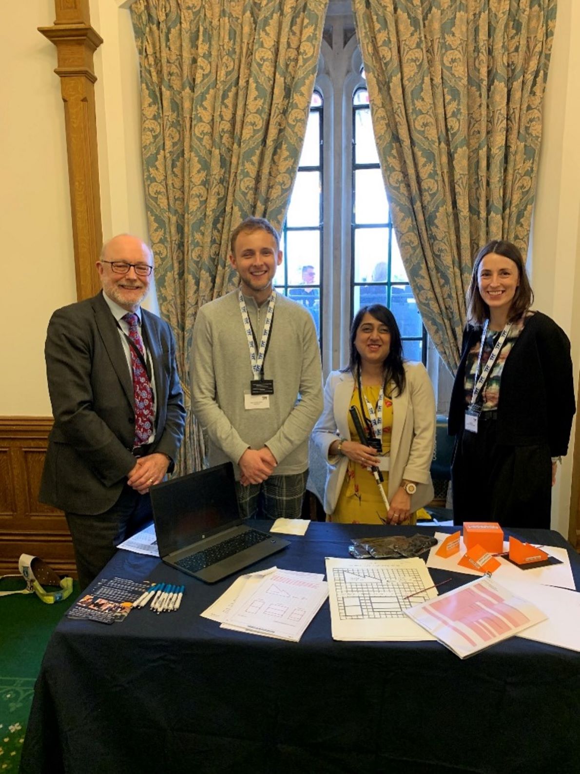 Aleks with an MP and TPT staff at a Can you See Me? event at Parliament standing smiling in front of some curtains and a table in front of them with papers on.