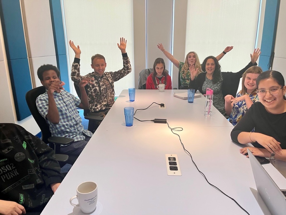 Aleks and other volunteers and staff celebrate in the TPT Hub around a table with arms raised in the air, having a great time.