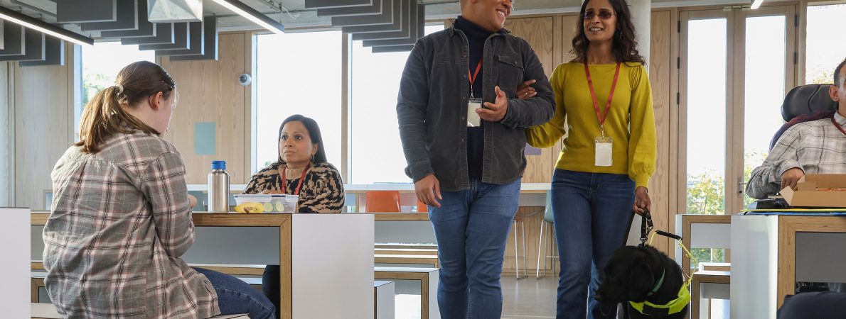 A man leads a blind or partially sighted woman through the canteen area of a workplace. They are smiling. The woman wears a yellow jumper and is holding the lead from her guide dog in her left hand. The man wears a grey denim jacket and has linked arms with the woman to guide her.