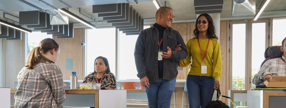 A man leads a blind or partially sighted woman through the canteen area of a workplace. They are smiling. The woman wears a yellow jumper and is holding the lead from her guide dog in her left hand. The man wears a grey denim jacket and has linked arms with the woman to guide her.