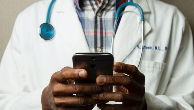 Photograph shows torso of a male doctor wearing a white coat with a stethoscope around his neck and holding a phone in both hands