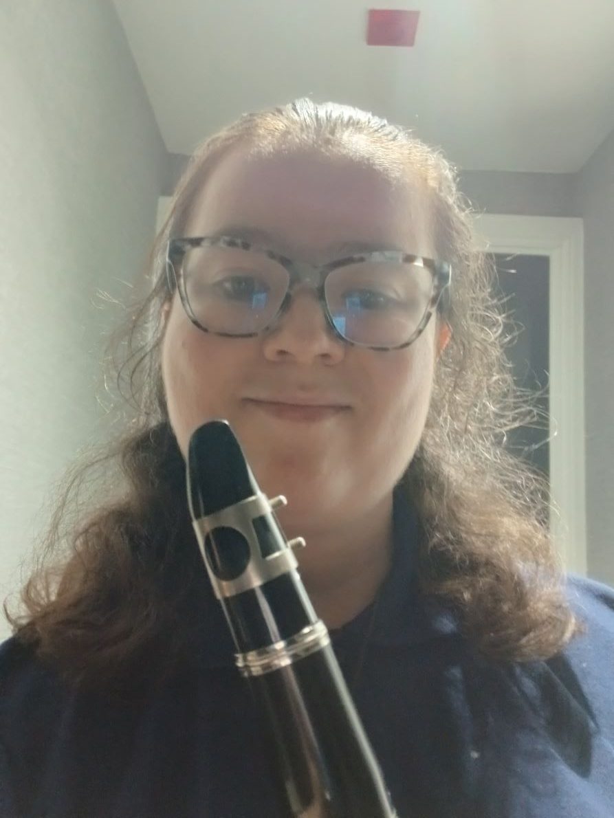 Sarah is wearing a purple shirt and holding her clarinet across her body, smiling at the camera.