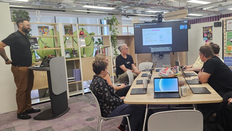 Image shows a man presenting to a group in an office setting. They are all looking towards the large screen at the end of the room while the man presenting is standing at a lectern.