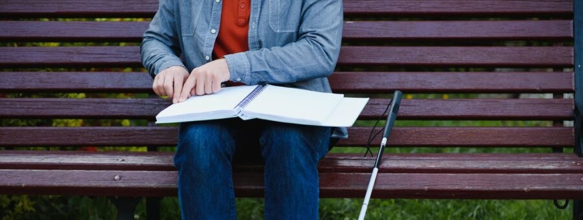 A student with a long cane is sitting on a bench reading a large open book in braille