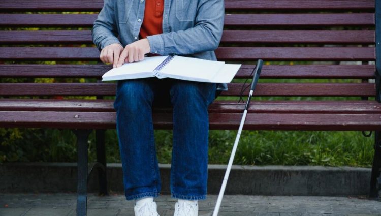 A student with a long cane is sitting on a bench reading a large open book in braille