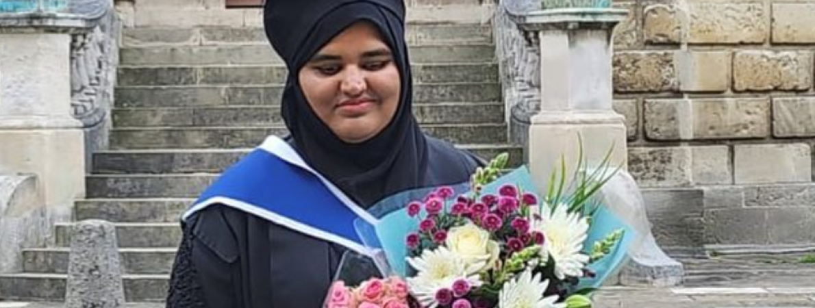 Khansa is standing outdoors on a stone walkway, wearing a black hijab, black graduation gown with a blue and white hood, and holding two colourful bouquets of flowers—one with pink roses and another with mixed white, yellow, and purple flowers.
