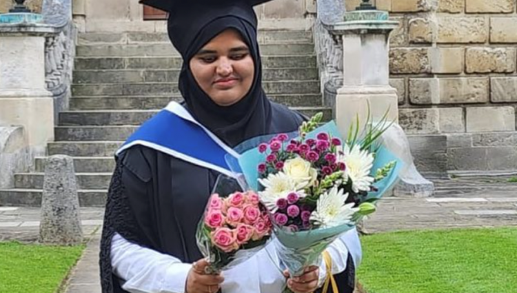 Khansa is standing outdoors on a stone walkway, wearing a black hijab, black graduation gown with a blue and white hood, and holding two colourful bouquets of flowers—one with pink roses and another with mixed white, yellow, and purple flowers.