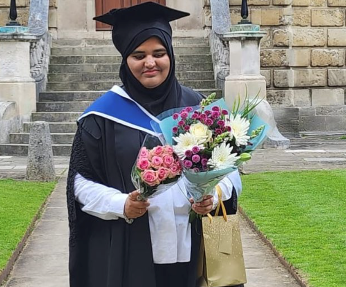 Khansa is standing outdoors on a stone walkway, wearing a black hijab, black graduation gown with a blue and white hood, and holding two colourful bouquets of flowers—one with pink roses and another with mixed white, yellow, and purple flowers.