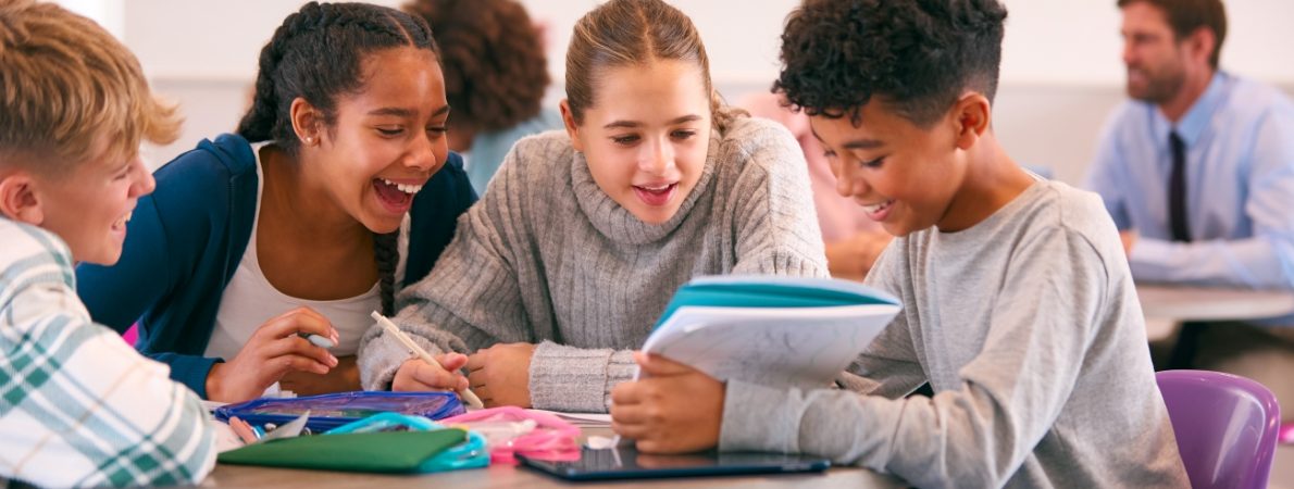 A group of schoolchildren gathered around a table in a classroom setting and talking to one another.