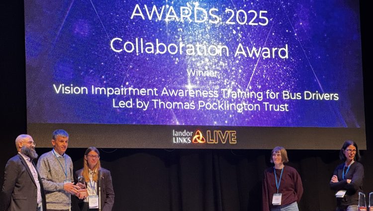 On stage at the award ceremony, a rather snazzy looking Dave and Lauren stand, holding their long canes next to a gentleman in a dark grey formal suit sporting a wonderfully bushy beard. A large screen behind them informs us of the celebrated winners with text that reads: Bus Centre of Excellence, Awards 2025, Collaboration Award, Winner, Vision Impairment Awareness Training for Bus Drivers Led by Thomas Pocklington Trust. The text is white on a sparkly blue background. The Screen behind is at least two Daves tall, and the stage must be at least four Daves wide, and the trio are standing to the left of the stage. To the right are two people who I am assuming are facilitators, one of whom is standing at a table draped in black cloth and the other standing looking towards the trio in awe. The lighting is an ambient blue with a soft spotlight on the winners.