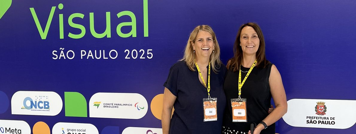 Emma and Cathy, holding a white cane, stand in front of the banner for the World Blindness Summit, São Paulo 2025, smiling and wearing event badges.