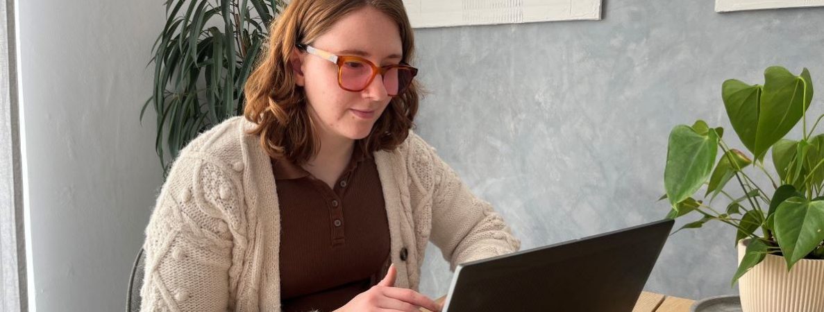 Chloe is sitting at a wooden table and is working on her laptop in a bright indoor space, with some potted plants on the table.