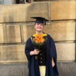 Sarah is wearing a navy graduation gown and cap, smiling at the camera and holding a bouquet of orange roses.