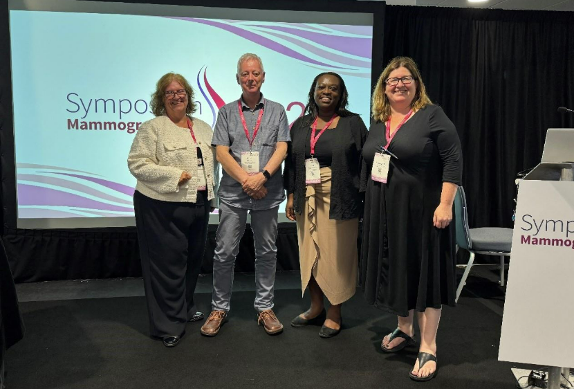 Alt text: A group of four people standing in front of a projector screen with the Symposium Mammographicum conference logo displayed.  From left to right we have-  Claire Borelli, Education Manager & Clinical Training Lead, St George’s National Breast Education Centre, Eamonn Dunne, Policy & Campaigns Manager at Thomas Pocklington Trust, Letitia Adamah-Miles,  Deputy Education Manager, St George’s National Breast Education Centre and Roisin Bradley, Director of breast screening at York and Scarborough Teaching Hospitals NHS Foundation Trust.