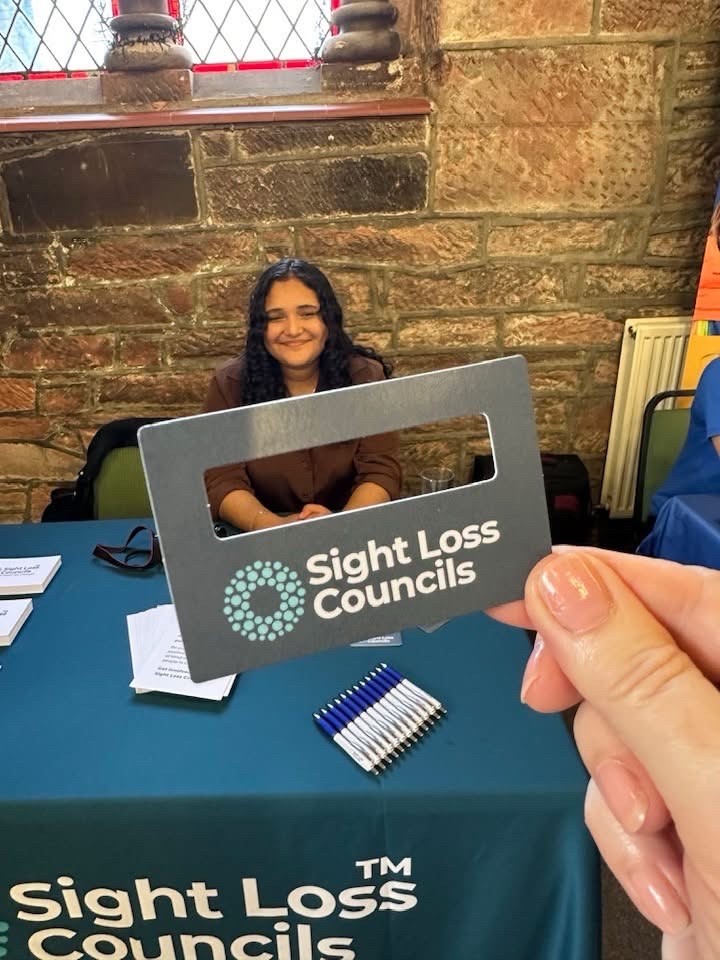 Pictured sitting behind the Sight Loss councils’ stall, Francesca Munoz, SLC Coordinator. The stall has brochures and merchandise, across the picture there is a hand holding the SLC’s signature guide. 