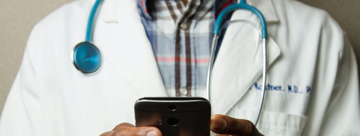 Photograph shows torso of a male doctor wearing a white coat with a stethoscope around his neck and holding a phone in both hands