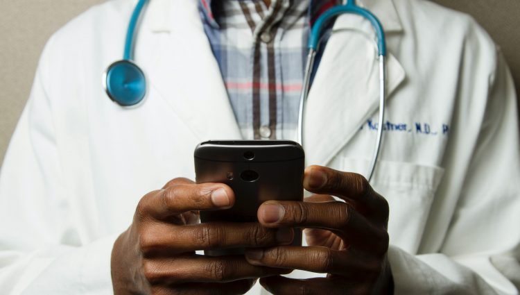 Photograph shows torso of a male doctor wearing a white coat with a stethoscope around his neck and holding a phone in both hands | links to article: Let’s Make Health Information Accessible