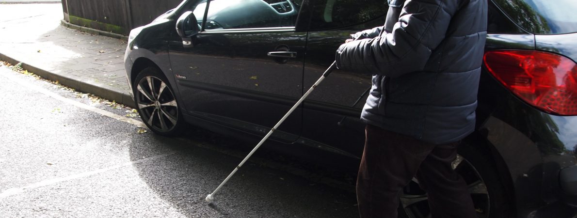Black hatchback vehicle is parked with 2 of its wheels on the pavement, blocking pedestrian access close to a fence. A visually impaired person using a long cane is on the main road trying to navigate a way past the vehicle.