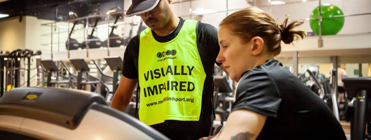 A woman showing a man with a vest that says 'Visually impaired' how to use the treadmill in a gym.