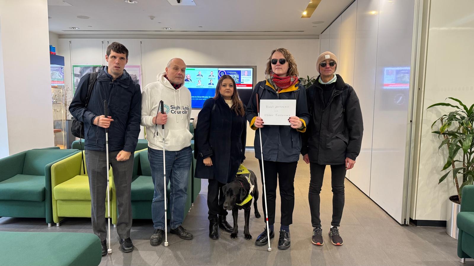 Five people, including Bhavini, standing indoors in the Department for Transport holding a card addressed to Heidi Alexander (Secretary of State for Transport) and Lilian Greenwood. They are also holding white canes and a guide dog is present.