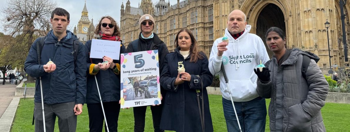 Group of campaigners standing outside the Houses of Parliament holding signs, cupcakes with candles and white canes, including a large birthday card that reads ‘5 long years of waiting’ with the TPT logo.