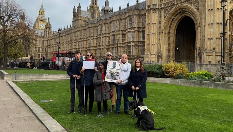Group of campaigners standing on grass outside the Houses of Parliament, holding white canes and a large birthday card that reads ‘5 long years of waiting’ with the TPT logo; a guide dog is also present