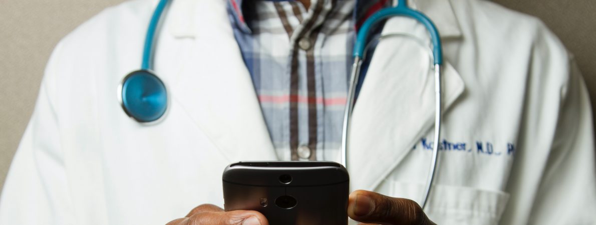 Photograph shows torso of a male doctor wearing a white coat with a stethoscope around his neck and holding a phone in both hands