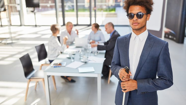 Man with a cane and dark glasses stands in a business meeting facing the camera. A desk with people in discussion is seen in the background.