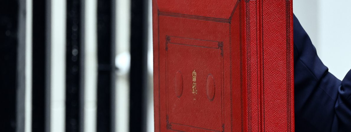 A close up of the Chancellor's Red Box held up by a hand with painted red nails.