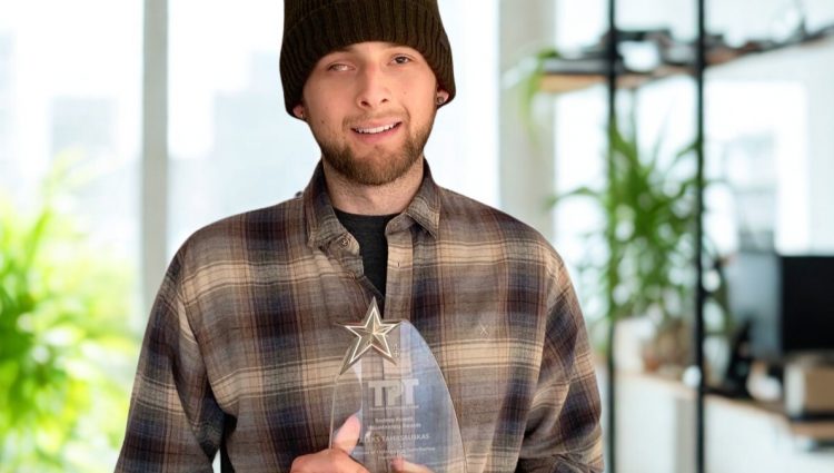 Aleks wears a brown checked shirt and a beanie, and smiles to the camera whilst holding a glass award.