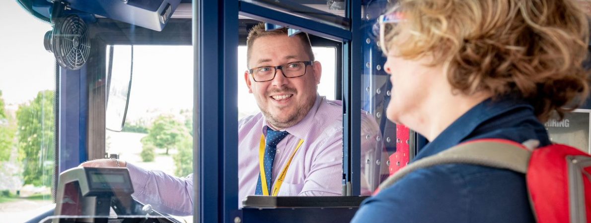 Male bus driver smiling as a passenger boards his bus. He is wearing glasses, a smart shirt and tie and has a yellow lanyard round his neck.