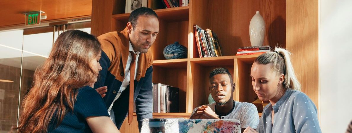 Four students sit studying around a table in a bright, modern university space. Two open laptops are on the table, behind them is a wooden bookshelf filled with books and decorative items.
